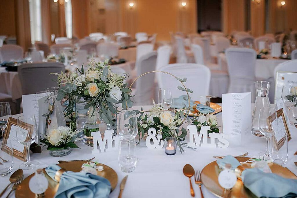 Ein mit weißem Leinen, goldenen Tellern, blauen Servietten und Grünzeug dekorierter Hochzeitstisch. Die Buchstaben "Mr. & Mrs." stehen in der Mitte zwischen Blumengestecken und Kerzen. Gläser, Speisekarten und gerahmte Fotos stehen auf dem Tisch; im Hintergrund sind weitere Tische zu sehen. Unser Zelt für Ihr Event in Leipzig und Umgebung.
