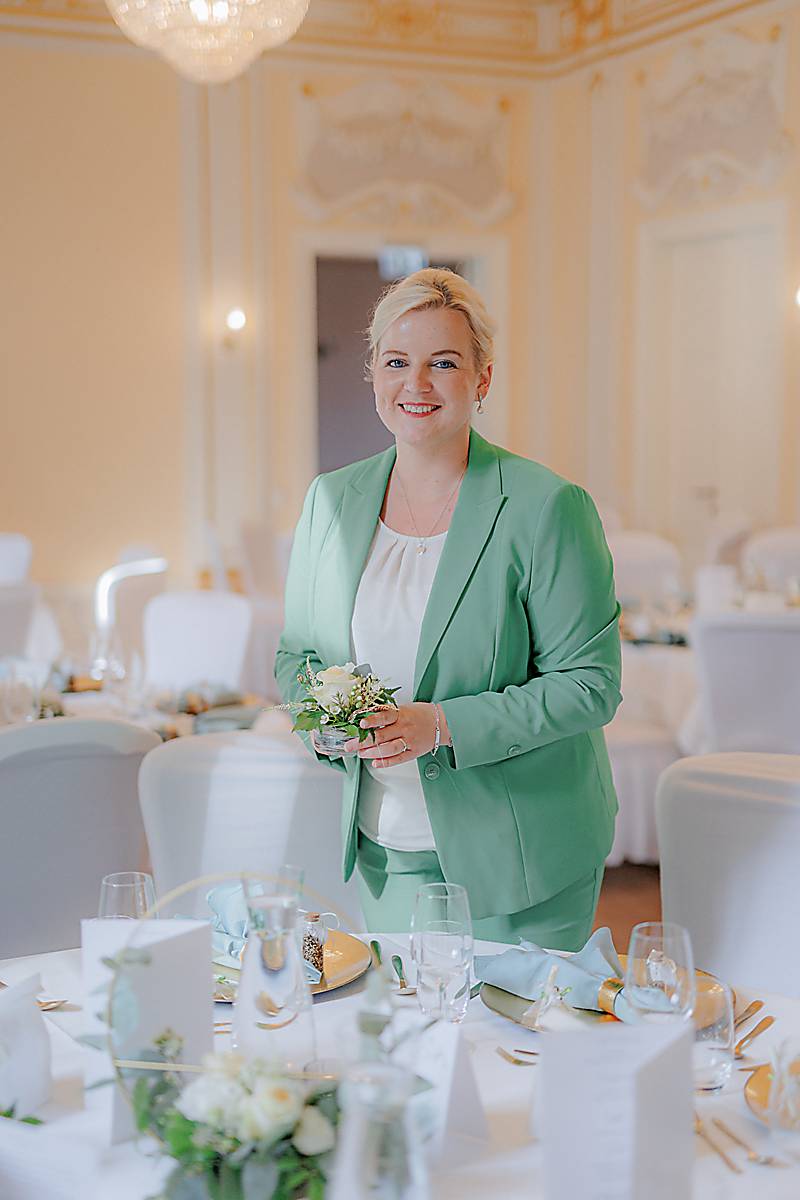 Eine Frau in einem hellgrünen Anzug hält ein kleines Blumenarrangement und lächelt, während sie in einem eleganten, sanft beleuchteten Speisesaal steht, der für eine Veranstaltung dekoriert ist. Die Tische sind mit weißem Leinen, Blumen, Gläsern und Speisekarten gedeckt. Die Atmosphäre ist formell und einladend. Unser Zelt für Ihr Event in Leipzig und Umgebung.