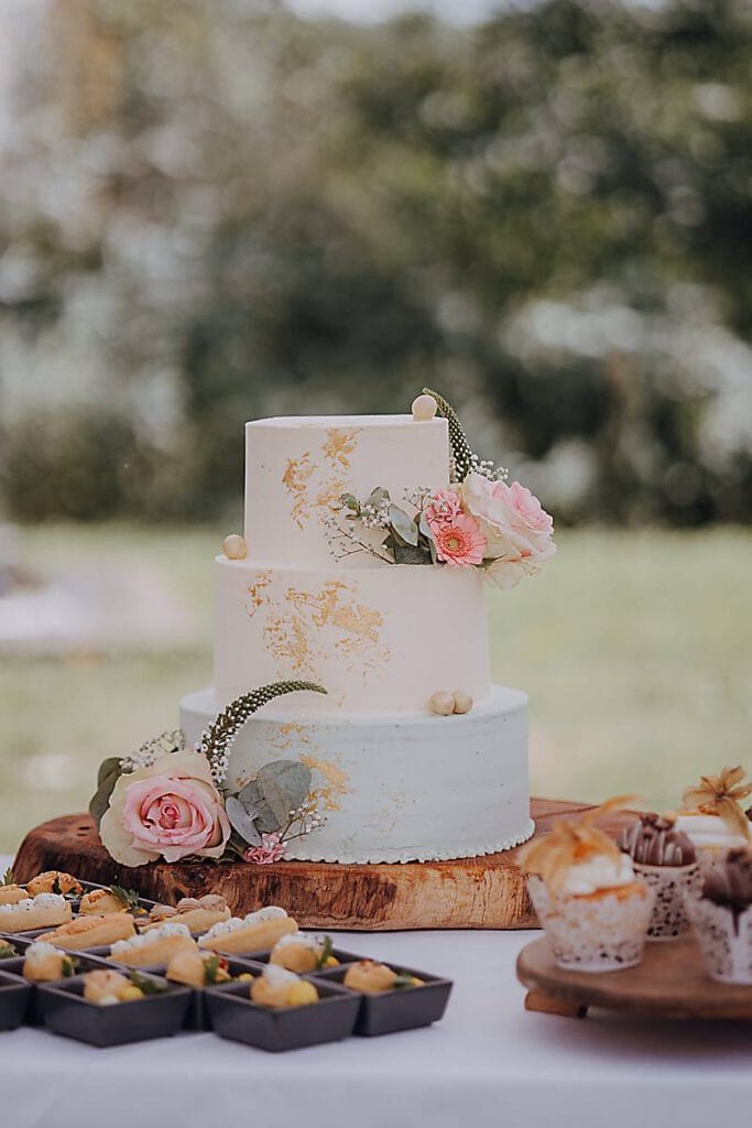 Eine wunderschöne zweistöckige Torte mit weißem und hellblauem Zuckerguss, verziert mit rosa Rosen, Grünzeug und goldenen Pinselstrichen, steht auf einem Holzständer auf Sarahs Hochzeit. Verschiedene mundgerechte Desserts umgeben sie auf einer weißen Tischdecke vor einem verschwommenen grünen Hintergrund. Unser Zelt für Ihr Event in Leipzig und Umgebung.