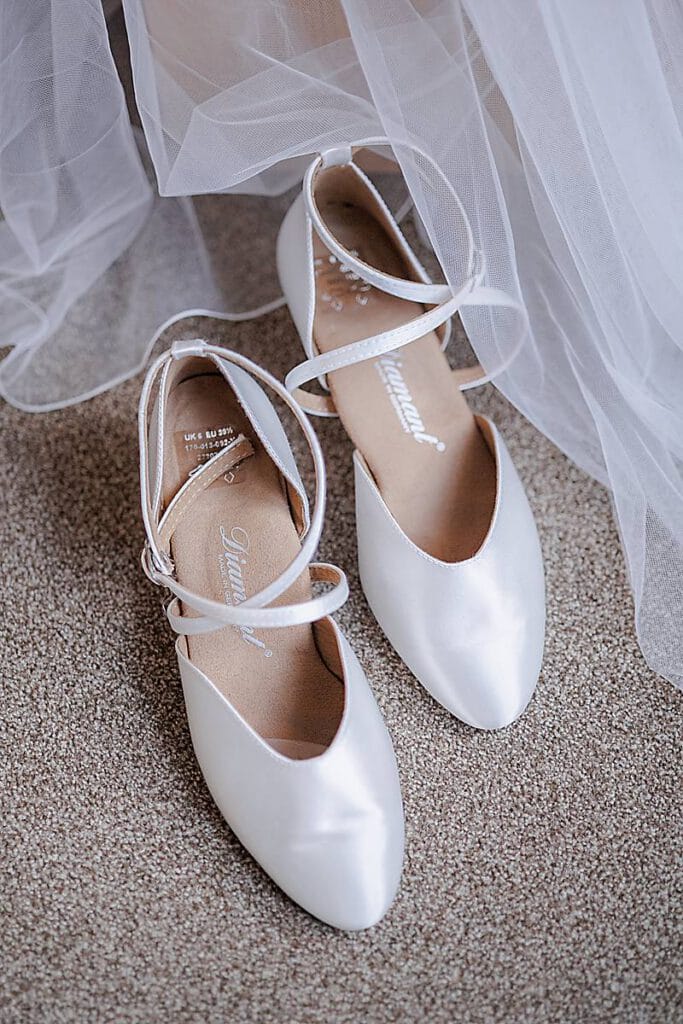 Ein Paar eleganter weißer Satin-Balletts mit überkreuzten Knöchelriemen steht auf einem beigen Teppich, der teilweise von weißem Tüll umrahmt ist, und erinnert an eine Braut, die mit Liebe und Herz erfüllt ist. Unser Zelt für Ihr Event in Leipzig und Umgebung.
