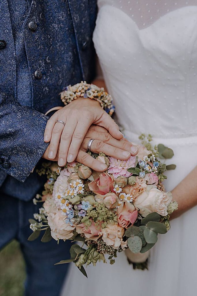 Nahaufnahme eines Paares in Hochzeitskleidung, das seine Hände mit den Eheringen auf einem Strauß pastellfarbener Blumen ruhen lässt. Die Braut trägt ein weißes Kleid mit durchsichtigem Tupfenüberzug, der Bräutigam einen blauen strukturierten Anzug - ein Moment für jede gute Seele. Unser Zelt für Ihr Event in Leipzig und Umgebung.
