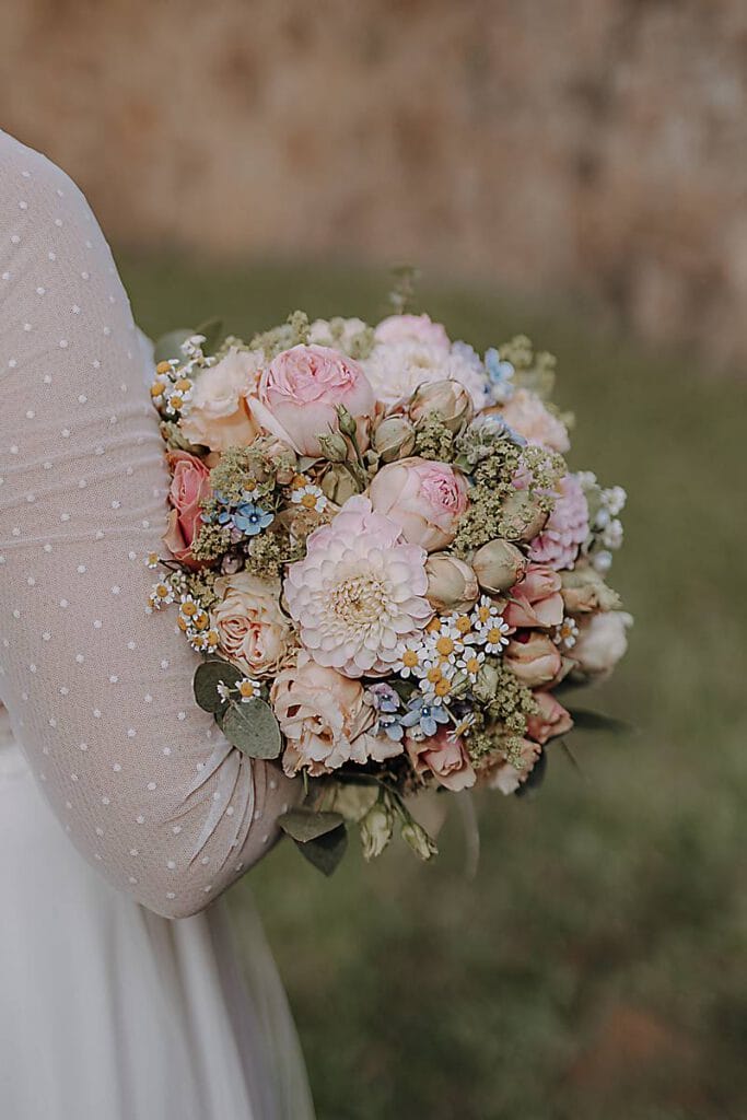 Eine Person in einem durchsichtigen, weißen, gepunkteten, langärmeligen Kleid hält einen Strauß mit pastellfarbenen Blumen. Das verschwommene grüne Gras und die Steinmauer bilden eine elegante Kulisse - eine ideale Inspiration für Dienstleister auswählen Tipps für Ihre nächste Veranstaltung. Unser Zelt für Ihr Event in Leipzig und Umgebung.