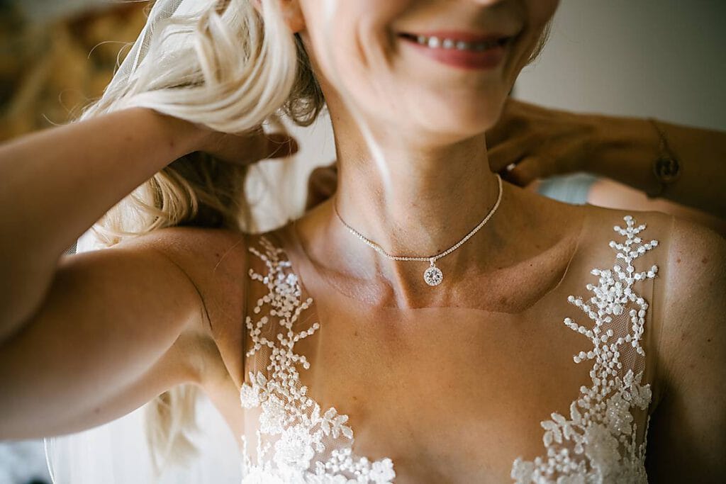 Eine lächelnde Braut rückt ihr Haar zurecht, während ihre beste Freundin ihr mit einer zarten Silberkette hilft. Sie trägt ein weißes Spitzenhochzeitskleid mit Blumendetails, während sanftes Sonnenlicht ihren freudigen Ausdruck und ihre elegante Kleidung hervorhebt. Unser Zelt für Ihr Event in Leipzig und Umgebung.
