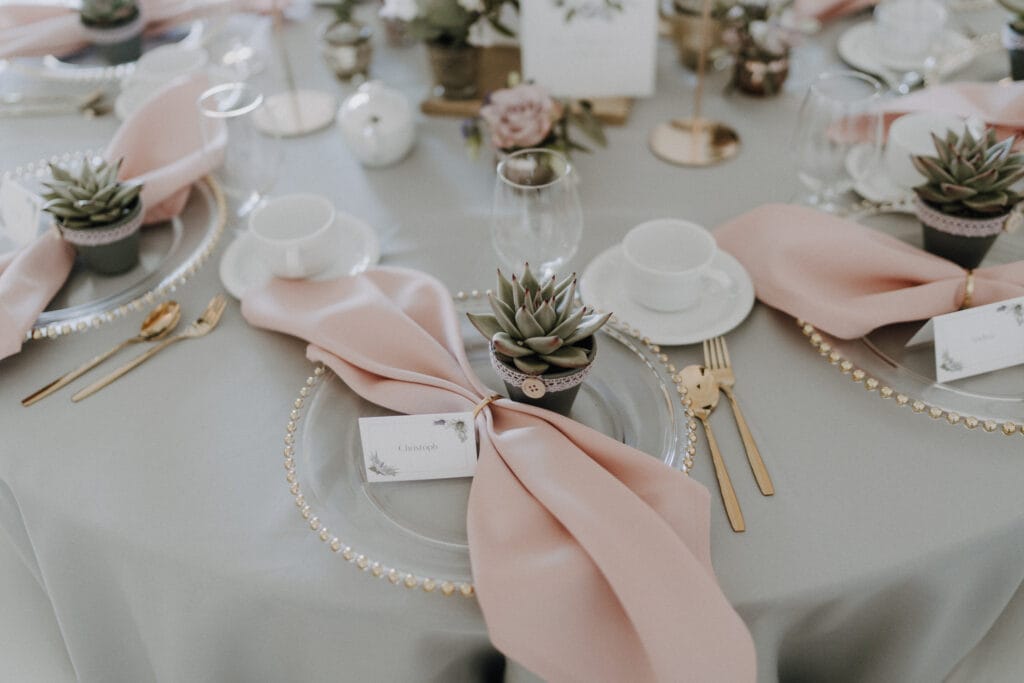 Ein runder Tisch, gedeckt für eine Veranstaltung mit einer grauen Tischdecke, goldenem Geschirr, durchsichtigen Tellern mit Perlenrand, rosa Servietten, personalisierten Namenskarten, kleinen getopften Sukkulenten als Gastgeschenke und eleganten Gläsern. Kerzen und Blumendekoration vervollständigen die einzigartige Szene. Unser Zelt für Ihr Event in Leipzig und Umgebung.