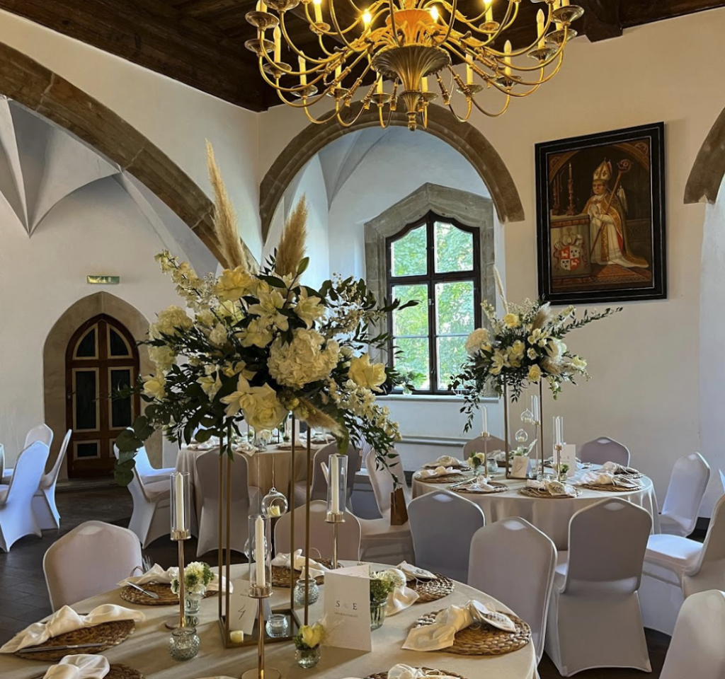 Ein heller, eleganter Speisesaal mit hohen Gewölbedecken und einem goldenen Kronleuchter - ideal, um Ihre Traumhochzeit zu feiern. Runde Tische mit weißen Tüchern, floralen Tafelaufsätzen und goldenen Akzenten, umgeben von weißen Stühlen, erwarten Sarah und ihre Gäste. Unser Zelt für Ihr Event in Leipzig und Umgebung.