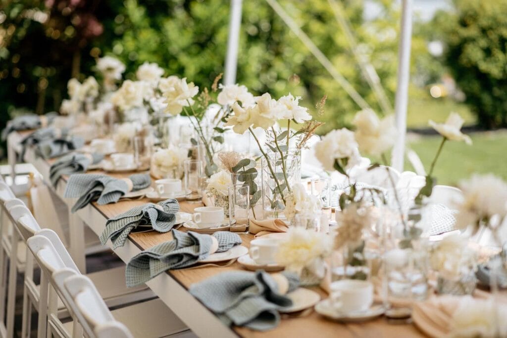 Ein langer Tisch im Freien ist elegant mit weißen Blumen, hellblauen Servietten und weißen Tassen gedeckt. Umgeben von weißen Stühlen und üppigem Grün, ist dieser Garten perfekt für Feiern - ideal, wenn Sie planen, Eventzelt mieten Leipzig oder brauchen eine Hochzeitsplanerin. Unser Zelt für Ihr Event in Leipzig und Umgebung.