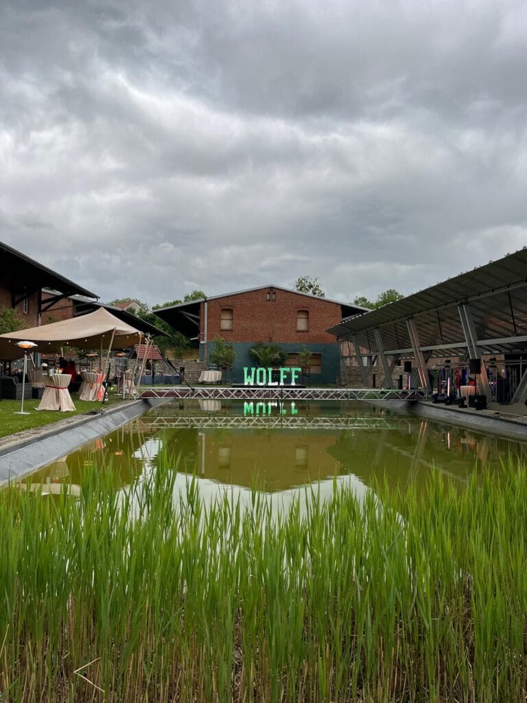 Ein rechteckiger, grünlicher Teich mit hohem Gras spiegelt ein Backsteingebäude und ein WOLFF MOTEL-Neonschild wider. Umgeben von Zelten und überdachten Gängen - perfekt für einen Aufbau unter freiem Himmel, wenn Sie auf der Suche nach einem Eventzelt mieten Leipzig sind oder eine Hochzeitsplanerin wie Sarah Schötter brauchen. Unser Zelt für Ihr Event in Leipzig und Umgebung.