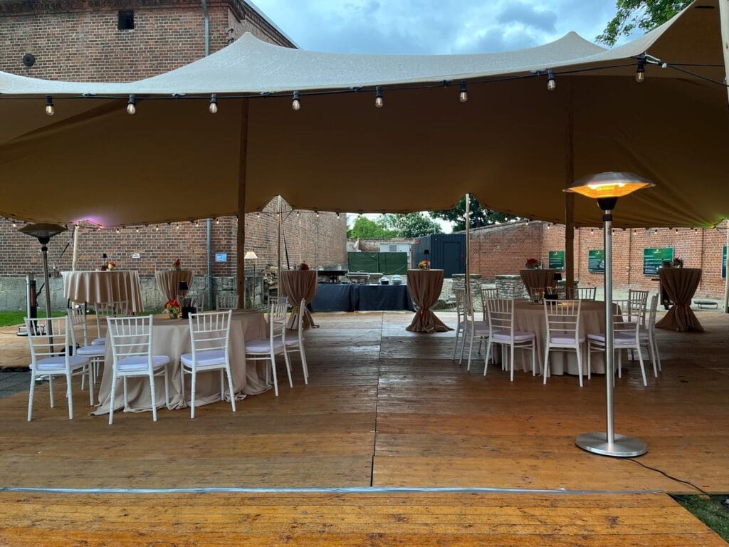 Event im Freien unter einem großen beigen Vordach mit Lichterketten, runden Tischen mit beigen Tüchern, weißen Stühlen und Stehtischen. Perfekt für Hochzeiten - lassen Sie Hochzeitsplanerin Sarah Schötter oder Eventzelt mieten Leipzig Ihren Tag zu etwas Besonderem machen!. Unser Zelt für Ihr Event in Leipzig und Umgebung.