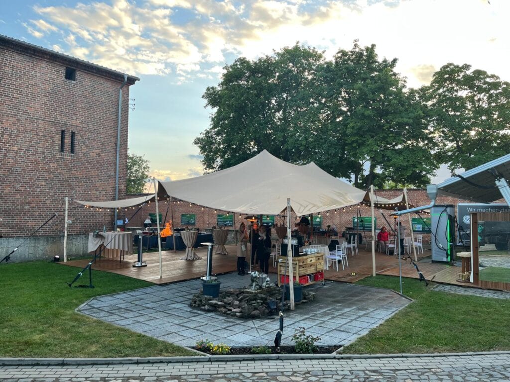Auf einem Holzdeck im Innenhof steht ein großes, beigefarbenes Zelt, das sich perfekt für das Eventzelt mieten Leipzig eignet. Umgeben von Backsteingebäuden und Grünflächen versammeln sich die Gäste unter dem Zelt an Tischen und Getränkestationen im sanften Licht des frühen Morgens oder späten Nachmittags. Unser Zelt für Ihr Event in Leipzig und Umgebung.