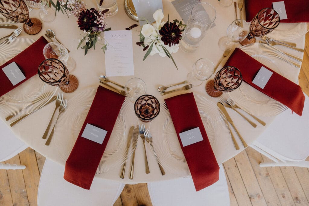 Ein runder Tisch für sechs Personen mit durchsichtigen Tellern, goldenem Besteck und burgunderroten Servietten, die jeweils mit einer Namenskarte versehen sind. Gläser im Vintage-Stil, ein floraler Mittelteil mit weißen und dunkelvioletten Blumen und eine Getränkekarte vervollständigen das elegante Arrangement. Unser Zelt für Ihr Event in Leipzig und Umgebung.