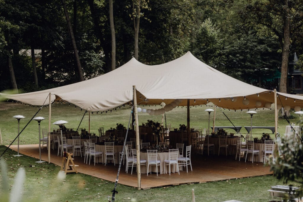 Ein großes beigefarbenes Zelt mit Lichterketten ist draußen auf einem grasbewachsenen Rasen aufgestellt. Unter dem Zelt sind runde Tische mit weißen Stühlen und floralen Tafelaufsätzen aufgestellt, bereit für eine Veranstaltung. Hohe Bäume und Grünpflanzen umgeben das Gelände und schaffen eine ruhige Atmosphäre. Unser Zelt für Ihr Event in Leipzig und Umgebung.