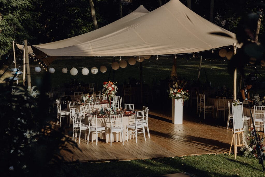 Ein Hochzeitsempfang unter einem beigen Vordachzelt in einem Garten. Runde Tische mit weißen Tischdecken, eleganten Blumenkränzen und weißen Stühlen sind auf einem Holzboden angeordnet. Hängende Papierlaternen und Lichterketten schmücken den Raum. Unser Zelt für Ihr Event in Leipzig und Umgebung.