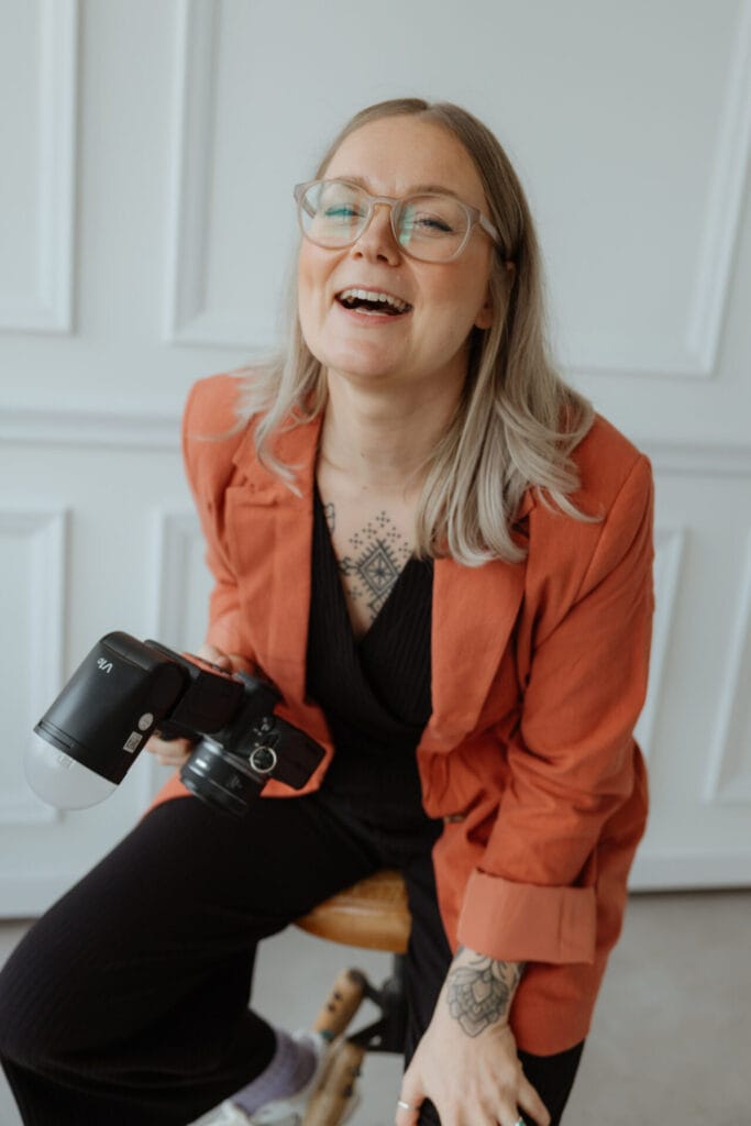 Elena Heymann, mit hellem Haar und Brille, lächelt, während sie eine Kamera hält und auf einem Hocker sitzt. Sie trägt einen orangefarbenen Blazer über einem schwarzen, der Tätowierungen auf ihrer Brust und ihrer Hand erkennen lässt. Die helle, moderne Umgebung ist mit weiß getäfelten Wänden ausgestattet. Unser Zelt für Ihr Event in Leipzig und Umgebung.